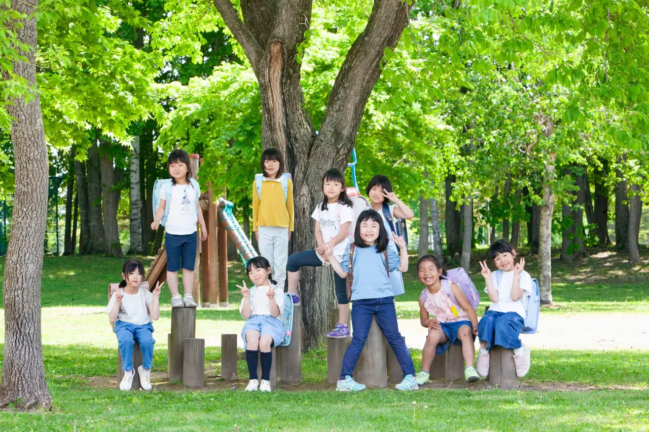 公園の大きな木の下で、ランドセルを背負った子どもたちが笑顔で集まっている様子